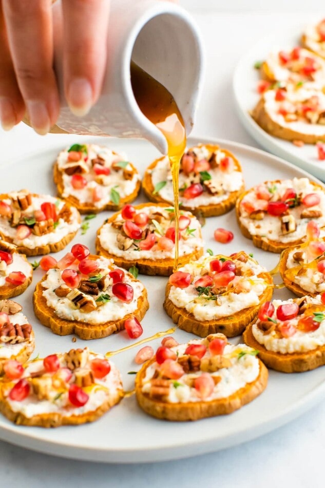 A hand is holding a small carafe of honey, drizzling the honey across a plate of sweet potato bites.