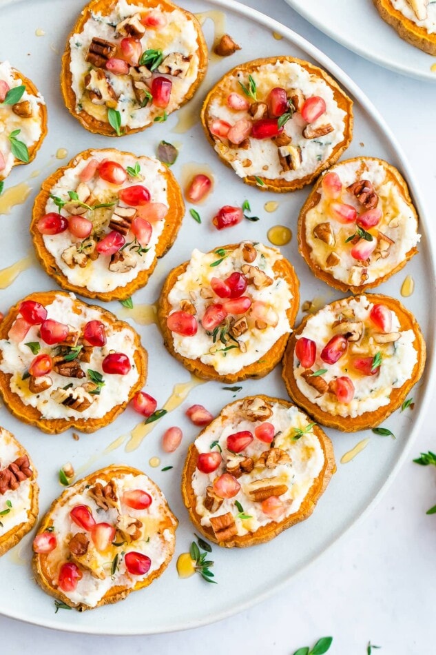 Overhead shot of a plate full of sweet potato bites topped with pomegranate arils, pecans, fresh thyme and drizzled with honey.