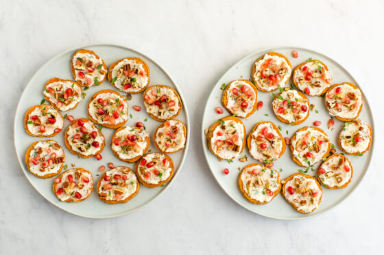 Two identical plates holding sweet potato rounds with whipped ricotta, pomegranate arils, pecans and fresh thyme sprinkled on top.