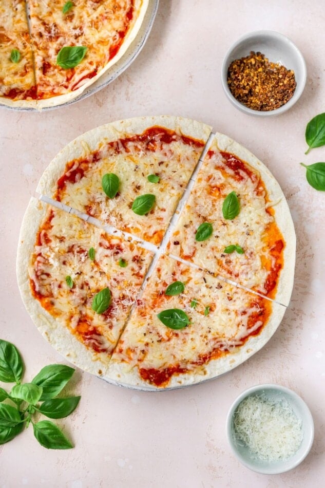 An overhead shot of a tortilla pizza with basil, cheese, and red pepper flakes in ramekins around the pizza.