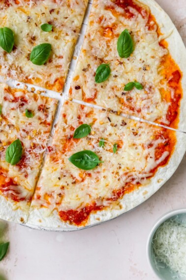 A close up overhead shot of a tortilla pizza. A ramekin of cheese is in the bottom right corner of the image.