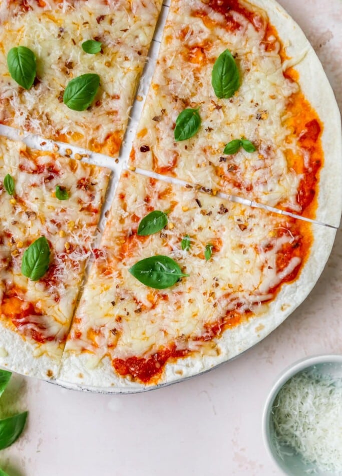 A close up overhead shot of a tortilla pizza. A ramekin of cheese is in the bottom right corner of the image.