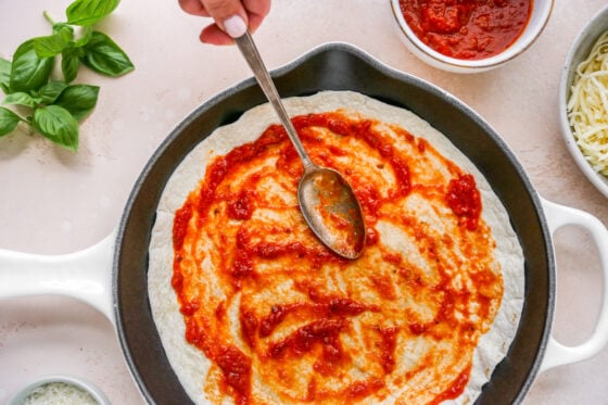 Pizza sauce spread around a tortilla with a spoon in a skillet.