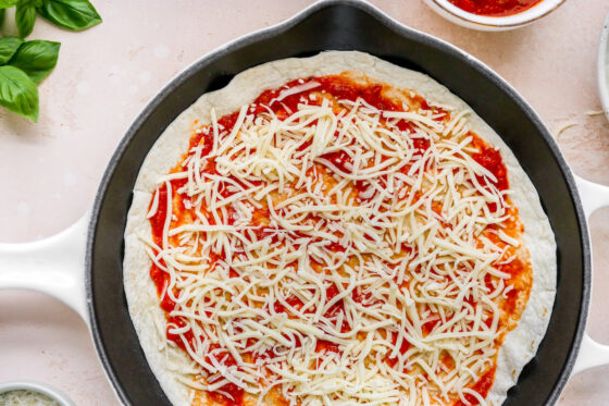 Cheese sprinkled onto of pizza sauce on a tortilla in a skillet.