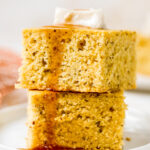 A closeup photo of two pieces of vegan cornbread stacked on top of each other drizzled with maple syrup and topped with a pat of vegan butter.