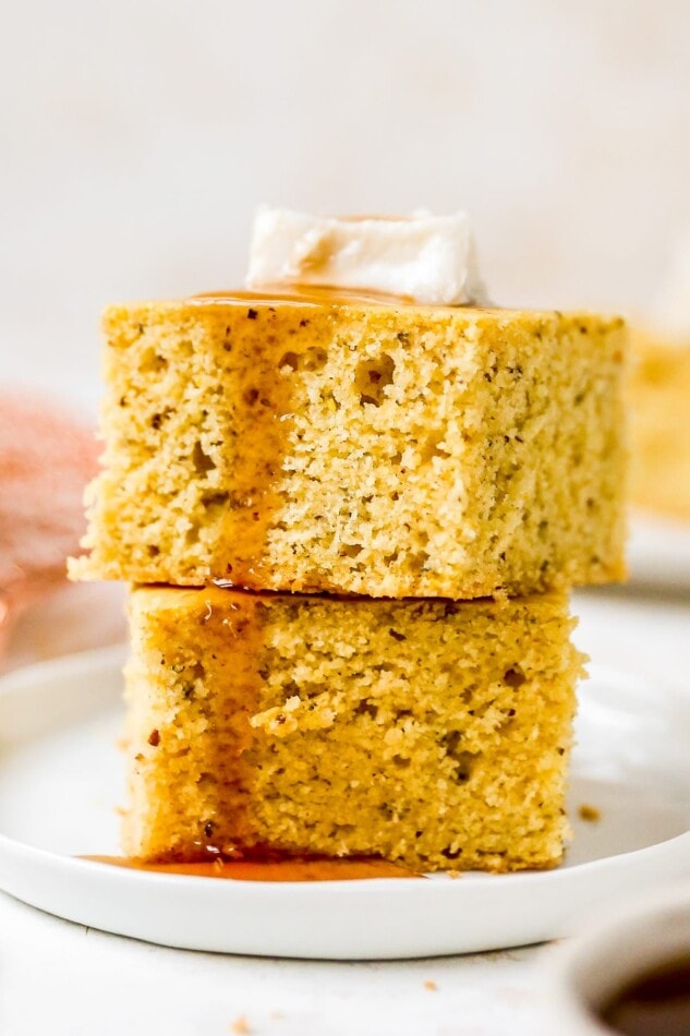 A closeup photo of two pieces of vegan cornbread stacked on top of each other drizzled with maple syrup and topped with a pat of vegan butter.