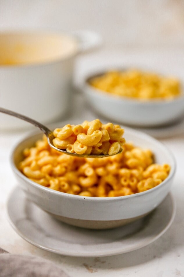A spoon with a bite of vegan mac and cheese being held up, with a bowl of mac and cheese out of focus in the background.