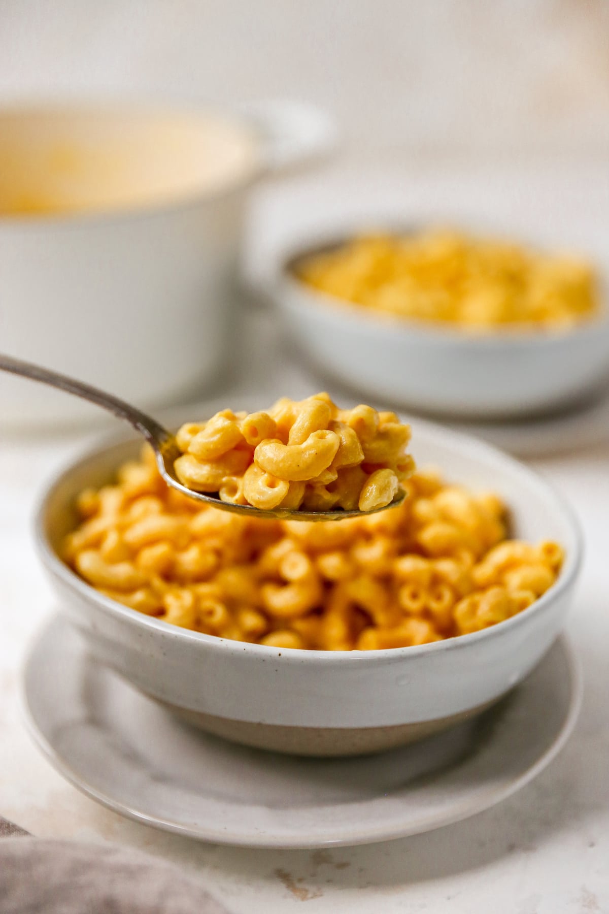 A spoon with a bite of vegan mac and cheese being held up, with a bowl of mac and cheese out of focus in the background.