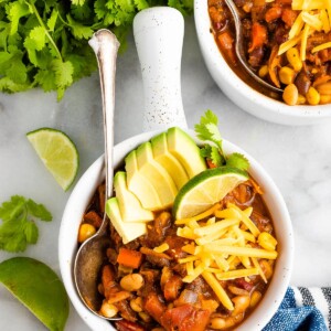 Overhead shot of a bowl of chili topped with avocado slices, cheese, and a lime wedge. A silver spoon rests inside the bowl. There are lime wedges and a bunch of cilantro around the bowl.