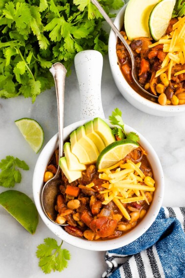 Overhead shot of a bowl of chili topped with avocado slices, cheese, and a lime wedge. A silver spoon rests inside the bowl. There are lime wedges and a bunch of cilantro around the bowl.