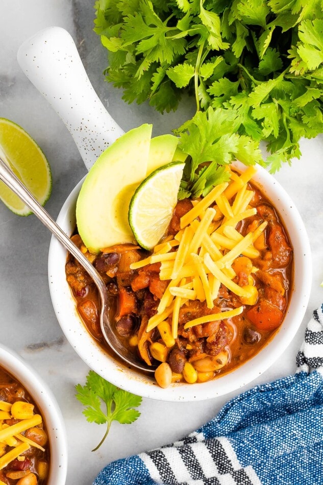 An overhead shot of a bowl of vegetarian chili, topped with avocado, shredded cheese, and a lime wedge.