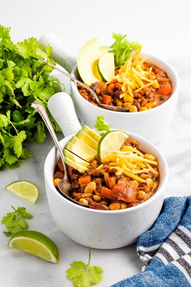 Two bowls of chili topped with avocado, lime wedge, and shredded cheese. A bunch of cilantro and some lime wedges are scattered around the bowls.