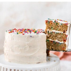 A cake sitting on a cake stand. The cake is iced in white frosting and topped with rainbow sprinkles. A slice has been cut and is being removed, exposing the 3 layers of funfetti cake with cream cheese frosting in between the layers.