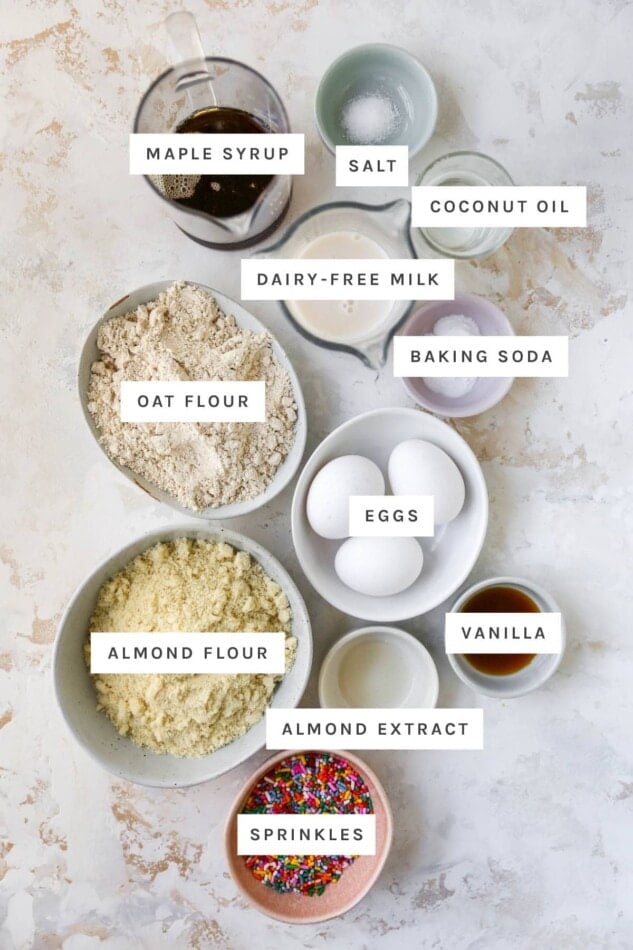 Ingredients in bowls measured out to make a healthy birthday cake: maple syrup, salt, coconut oil, dairy-free milk, baking soda, oat flour, eggs, vanilla, almond flour, almond extract and sprinkles.