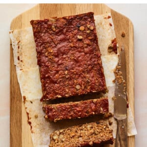An overhead shot of a cutting board with a partially sliced lentil loaf on parchment paper. A knife lays on the cutting board perpendicular to the loaf.