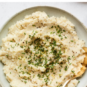 A bowl of mashed cauliflower topped with chives. A serving spoon rests inside the bowl.