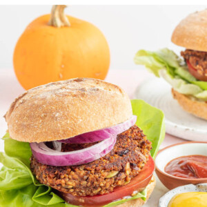 A black bean pumpkin burger on a bun with lettuce, tomato and onion. The burger is on a plate with two small ramekins of mustard and ketchup. There is a small pumpkin in the background.