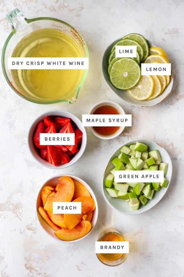 Ingredients measured out to make white wine sangria: dry white wine, lime, lemon, maple syrup, berries, green apple, peach and brandy.