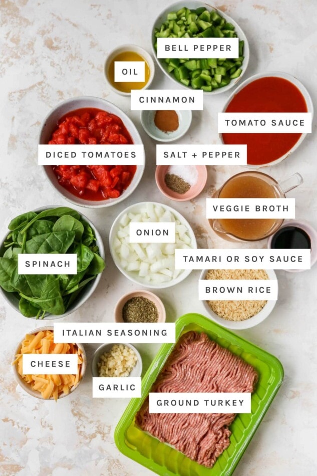 Ingredients measured out in bowls to make stuffed pepper casserole: chopped bell pepper, oil, cinnamon, tomato sauce, salt, pepper, diced tomatoes, veggie broth, onion, soy sauce, spinach, brown rice, italian seasoning, garlic, cheese and ground turkey.