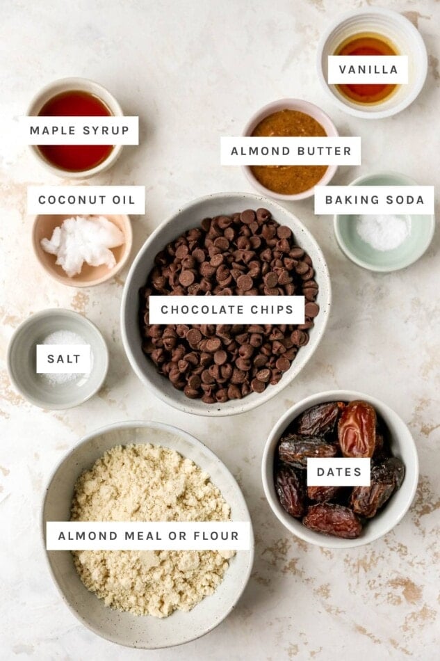 Ingredients measured out to make healthy Twix bars: vanilla, maple syrup, almond butter, coconut oil, baking soda, chocolate chips, salt, dates and almond meal/flour.