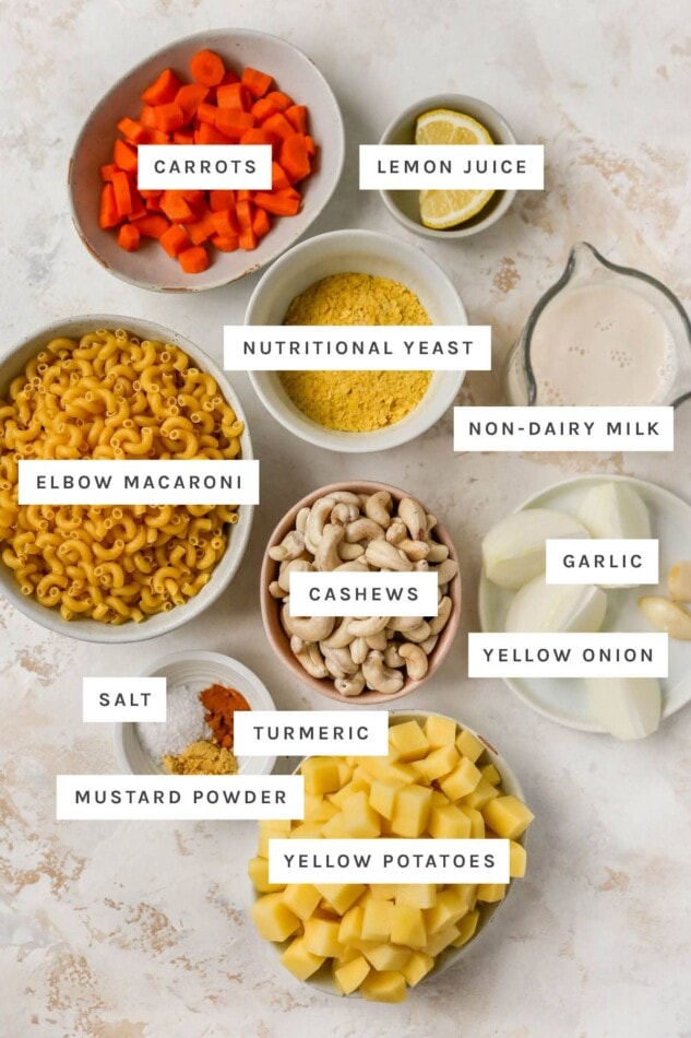 Ingredients measured out in bowls and on plates to make vegan mac and cheese: carrots, lemon, nutritional yeast, elbow macaroni, non-dairy milk, cashews, garlic, onion, salt, turmeric, mustard powder and potatoes.