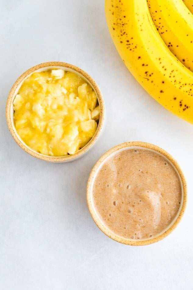 An overhead photo of two small dishes, one with mashed banana and one with banana puree. Whole bananas are in the top corner.