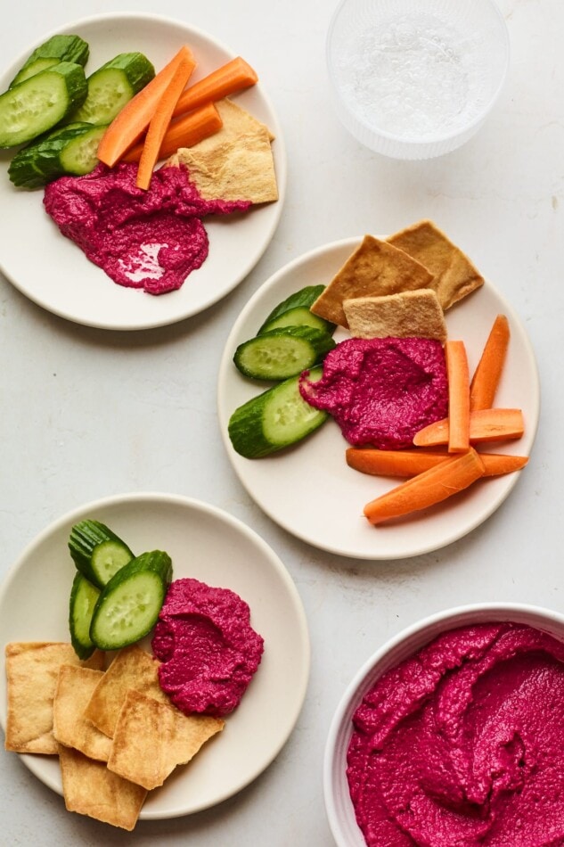 3 plates with a scoop of beet hummus and various vegetables for dipping. A bowl of beet hummus is in the bottom right corner.