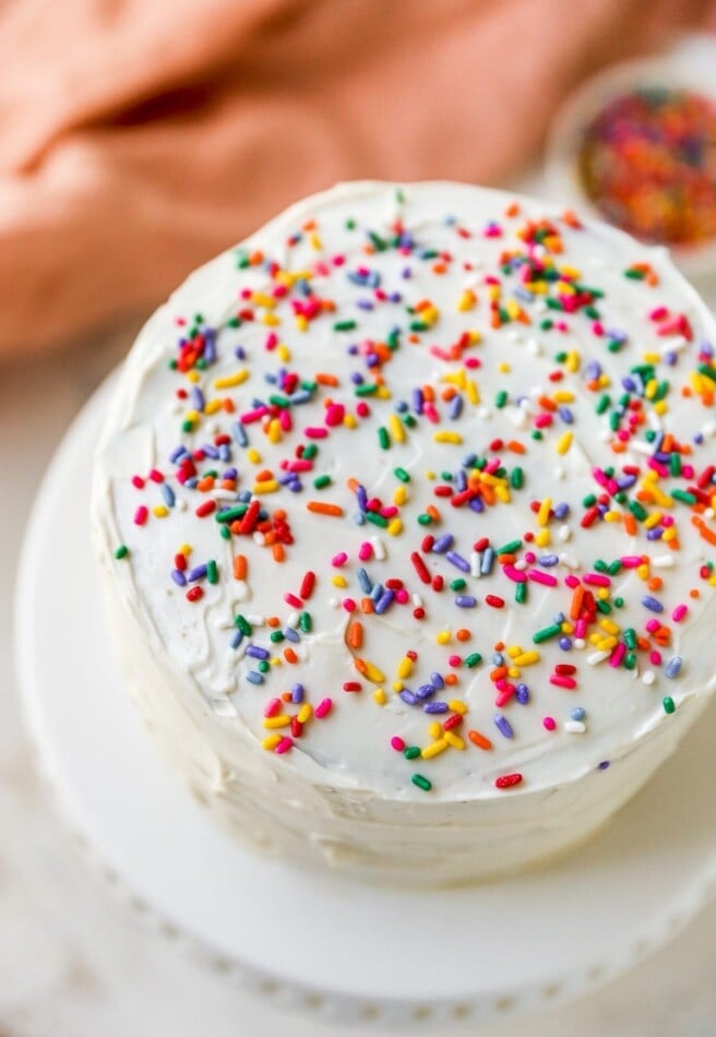 A close up photo of a cake sitting on a cake stand. The cake is iced in white frosting and topped with rainbow sprinkles.