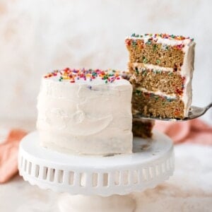 A cake sitting on a cake stand. The cake is iced in white frosting and topped with rainbow sprinkles. A slice has been cut and is being removed, exposing the 3 layers of funfetti cake with cream cheese frosting in between the layers.