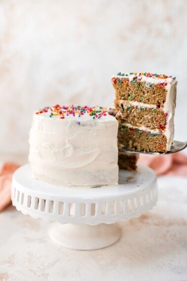 A cake sitting on a cake stand. The cake is iced in white frosting and topped with rainbow sprinkles. A slice has been cut and is being removed, exposing the 3 layers of funfetti cake with cream cheese frosting in between the layers.