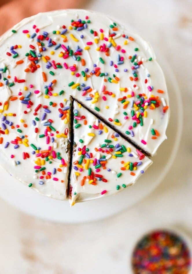 Overhead photo of a cake on a cake stand. The cake is iced in white frosting and topped with rainbow sprinkles. A slice has been cut but not removed.