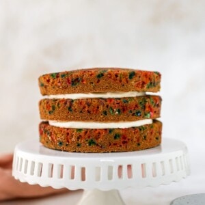 A white cake stand with 3 layers of birthday cake stacked on top. There is cream cheese icing between the layers.