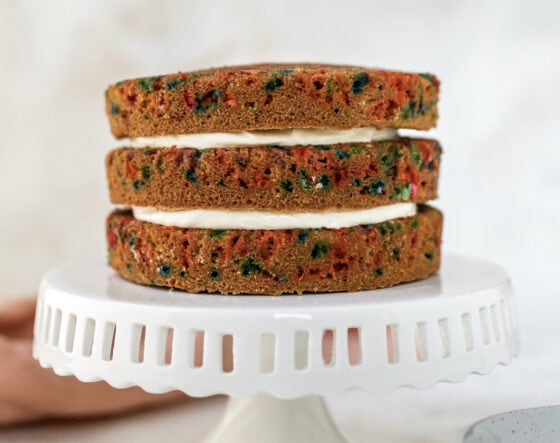 A white cake stand with 3 layers of birthday cake stacked on top. There is cream cheese icing between the layers.