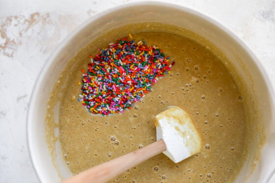 Cake batter in a bowl. A spatula rests in the bowl alongside some rainbow sprinkles that have yet to be mixed in.