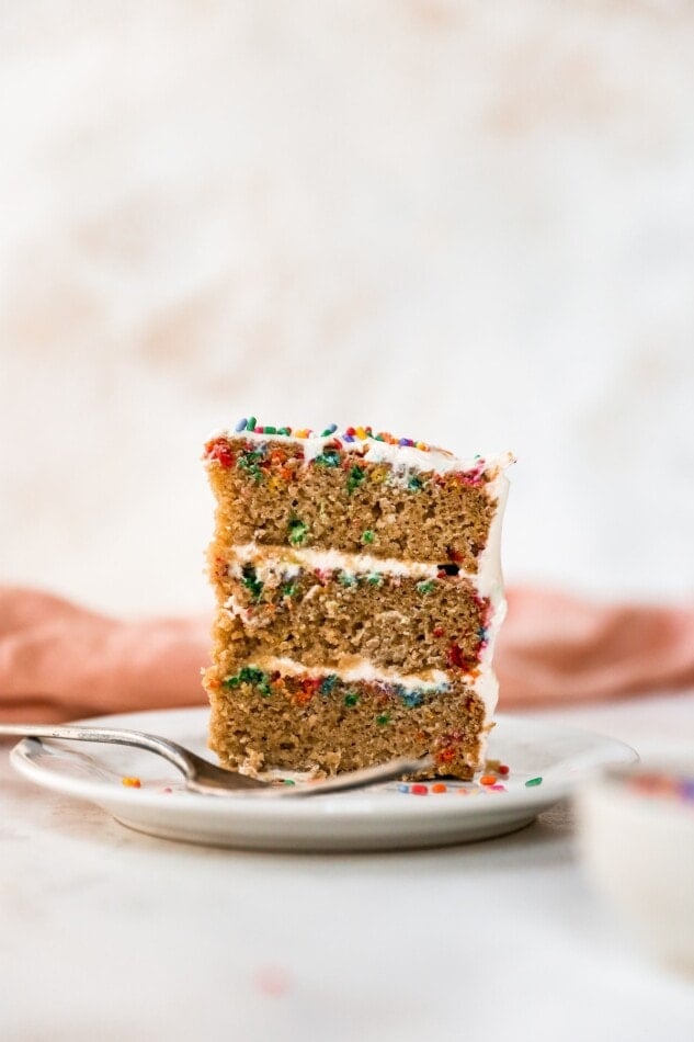 A 3-layer slice of healthy birthday cake with layers of cream cheese icing and topped with rainbow sprinkles sits on a plate with a fork.