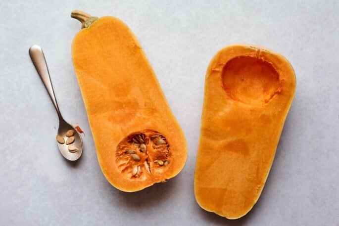 A butternut squash cut in half. One half has the seeds scooped out, the other still has the seeds.