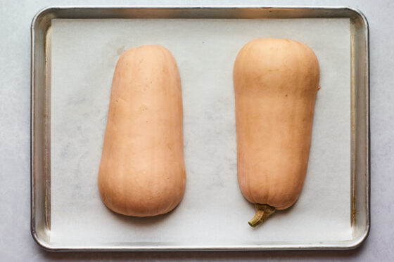 Two butternut squash halves on a sheet of parchment paper on a sheet pan.