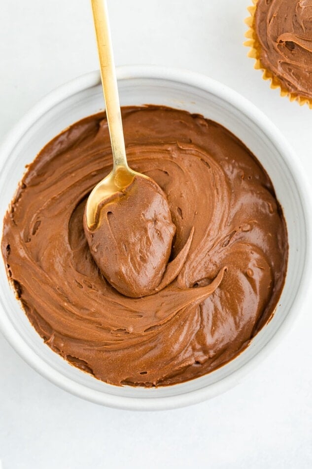 An overhead photo of a bowl of health chocolate frosting. A gold spoon rests inside of the bowl with a scoop of frosting on top of it.