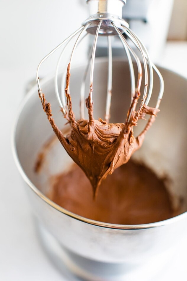 A stand-mixer whisk attachment with chocolate frosting on it. The bowl is out of focus in the background with additional chocolate frosting.