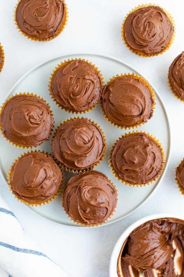 An overhead photo of a plate of cupcakes that have been topped with chocolate frosting. Additional cupcakes are scattered around the plate.