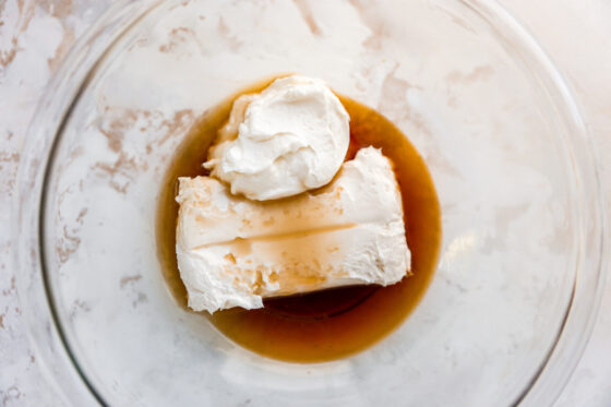 A glass mixing bowl with ingredients for cream cheese frosting: cream cheese, greek yogurt, vanilla extract and maple syrup.