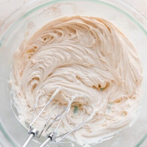 A glass mixing bowl with ingredients for cream cheese frosting that have been whipped together. Two mixers from a handheld mixer are resting inside the bowl.