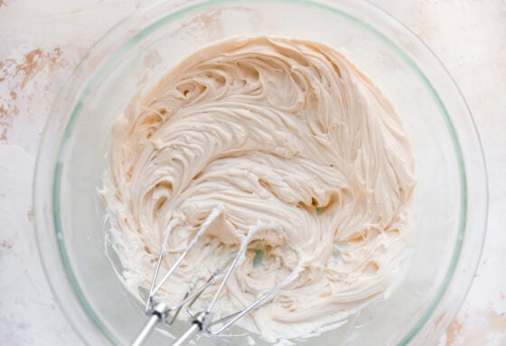 A glass mixing bowl with ingredients for cream cheese frosting that have been whipped together. Two mixers from a handheld mixer are resting inside the bowl.