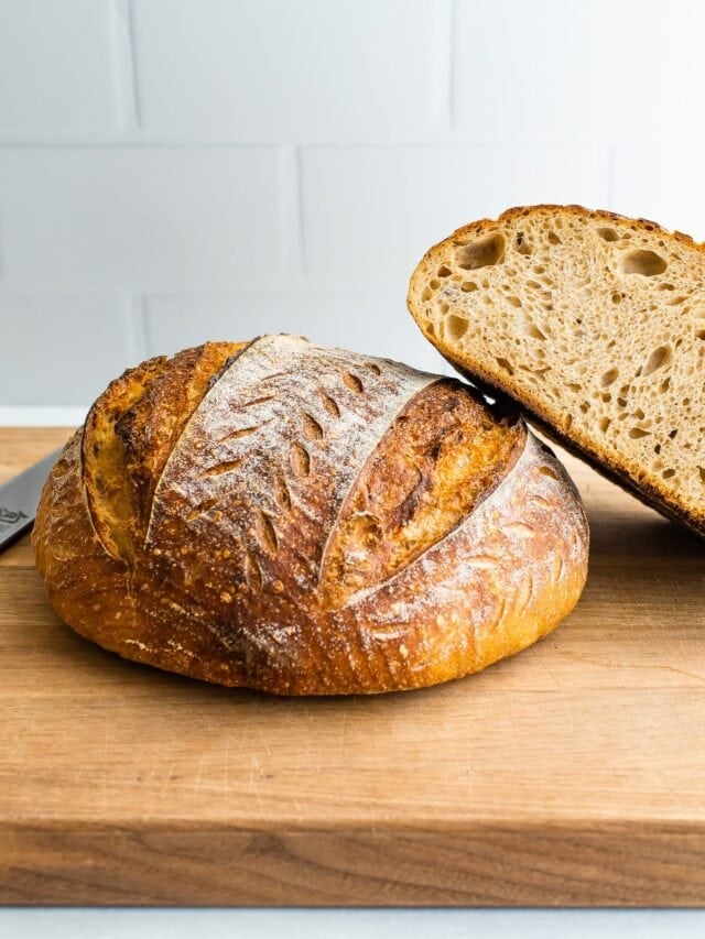 A loaf of sourdough bread on a wooden cutting board with a loaf that has been cut in half leaning against it.