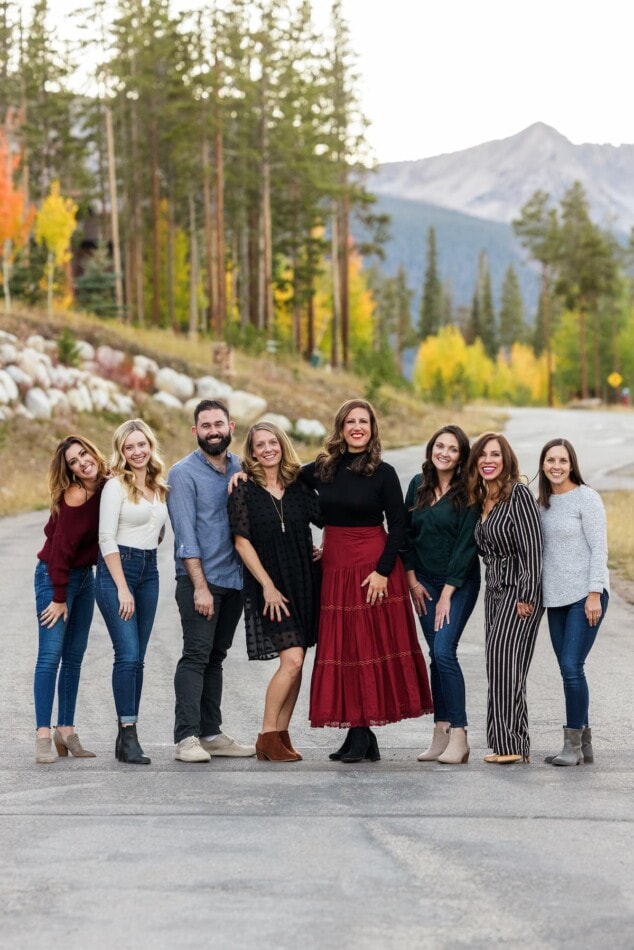 8 people lined up in a row smiling at the camera. A mountain and trees are in the backdrop.