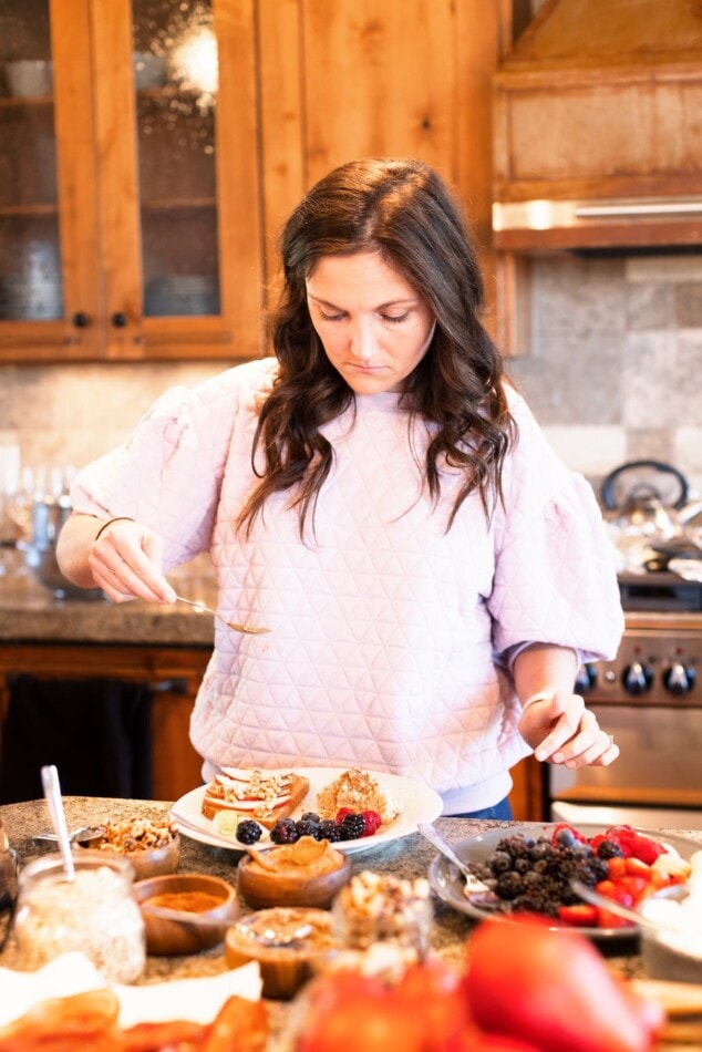A woman with long brown hair is sprinkling an unidentified item on top of a plate with berries and toast on top. There are several plates around the countertop holding various breakfast foods and toppings.