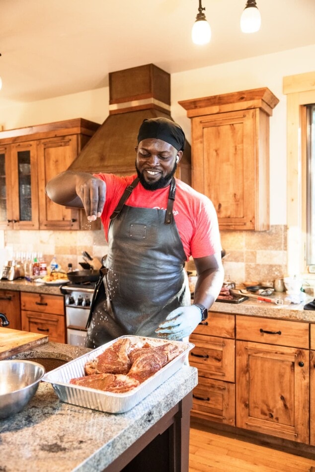 A photo of a man smiling, wearing a black apron and chefs hat, seasoning meat in a tinfoil disposable pan.