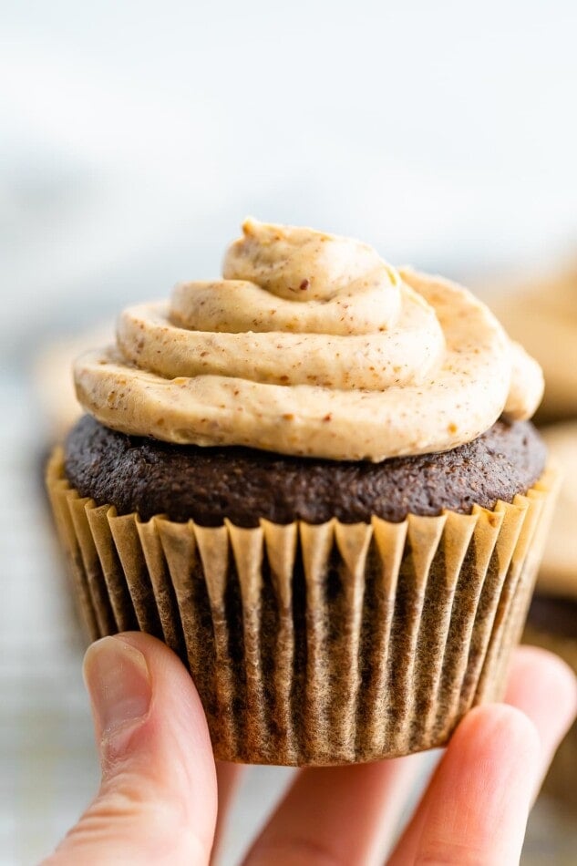 A chocolate cupcake in a brown wrapper topped with healthy greek yogurt frosting. The cupcake is being held up by a hand.