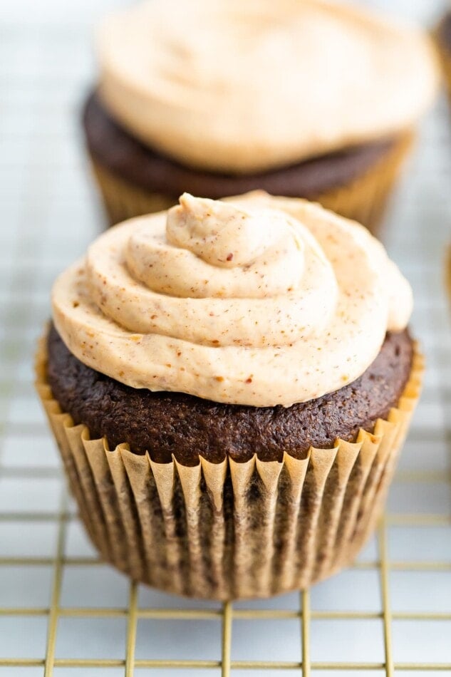 A chocolate cupcake in a brown wrapper topped with healthy greek yogurt frosting. The cupcake is resting on a gold wire rack.