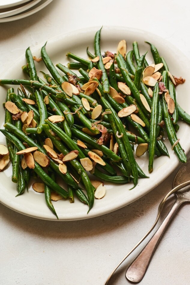 An closeup overhead photo of a serving dish holding green beans almondine. A serving fork and spoon lay next to the dish.